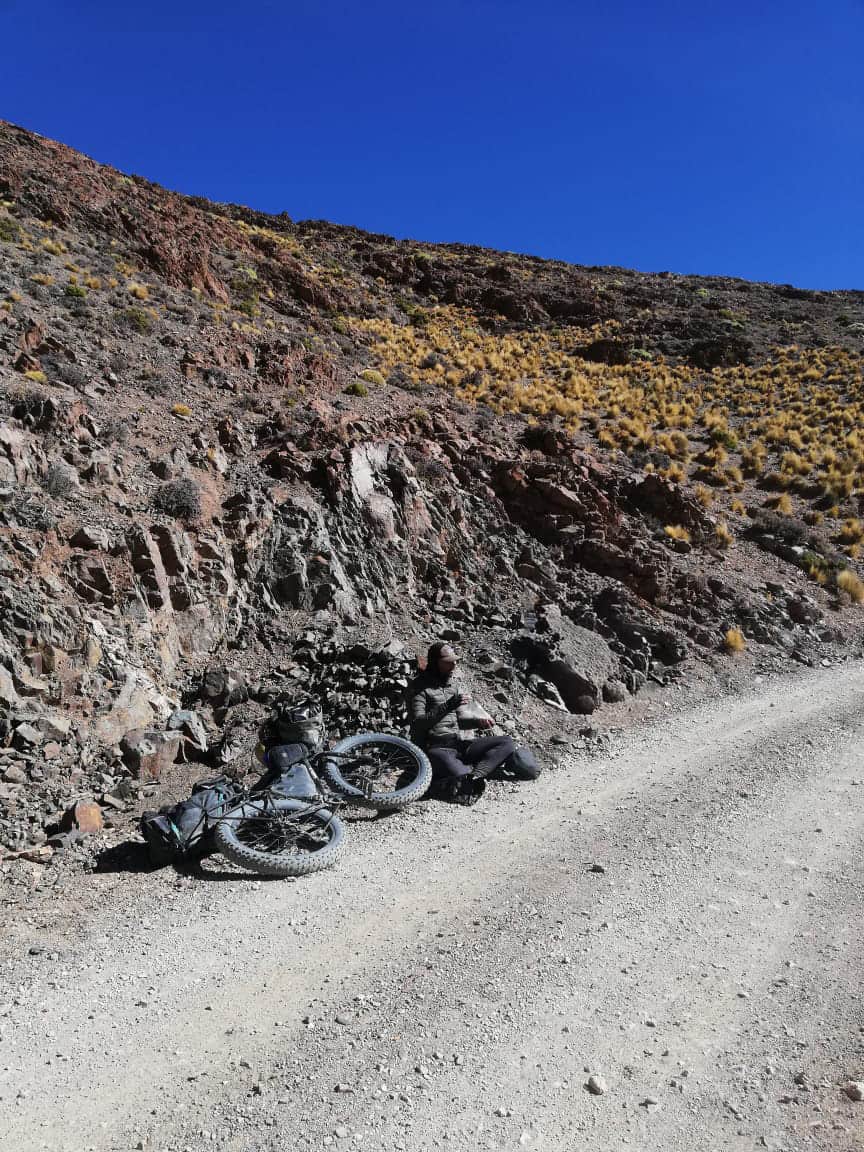 Cyclist resting beside a loaded touring bike on a gravel mountain road
